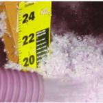 A pink hose blowing in fiberglass insulation with a measuring stick showing its depth at 20 inches.