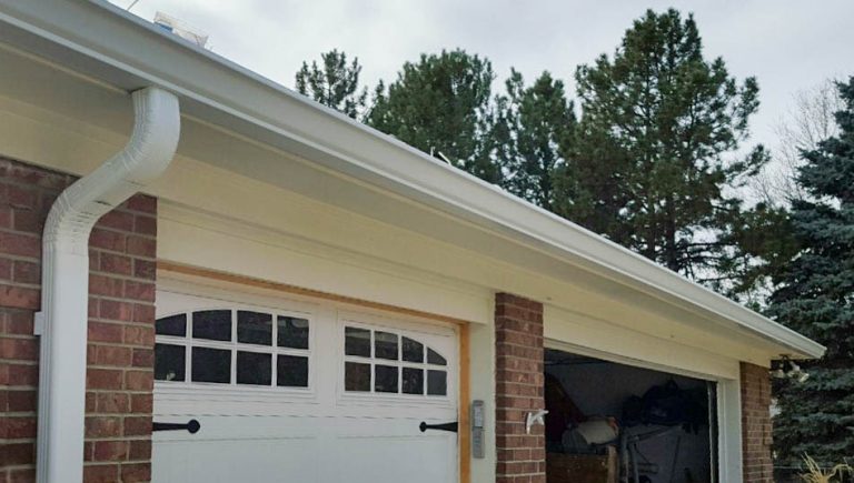 White rain gutters on a home placed above the garage. A drain pipe is seen going to the ground on one side. The left garage door is white and has sectioned windows at the top. The right garage door is open.
