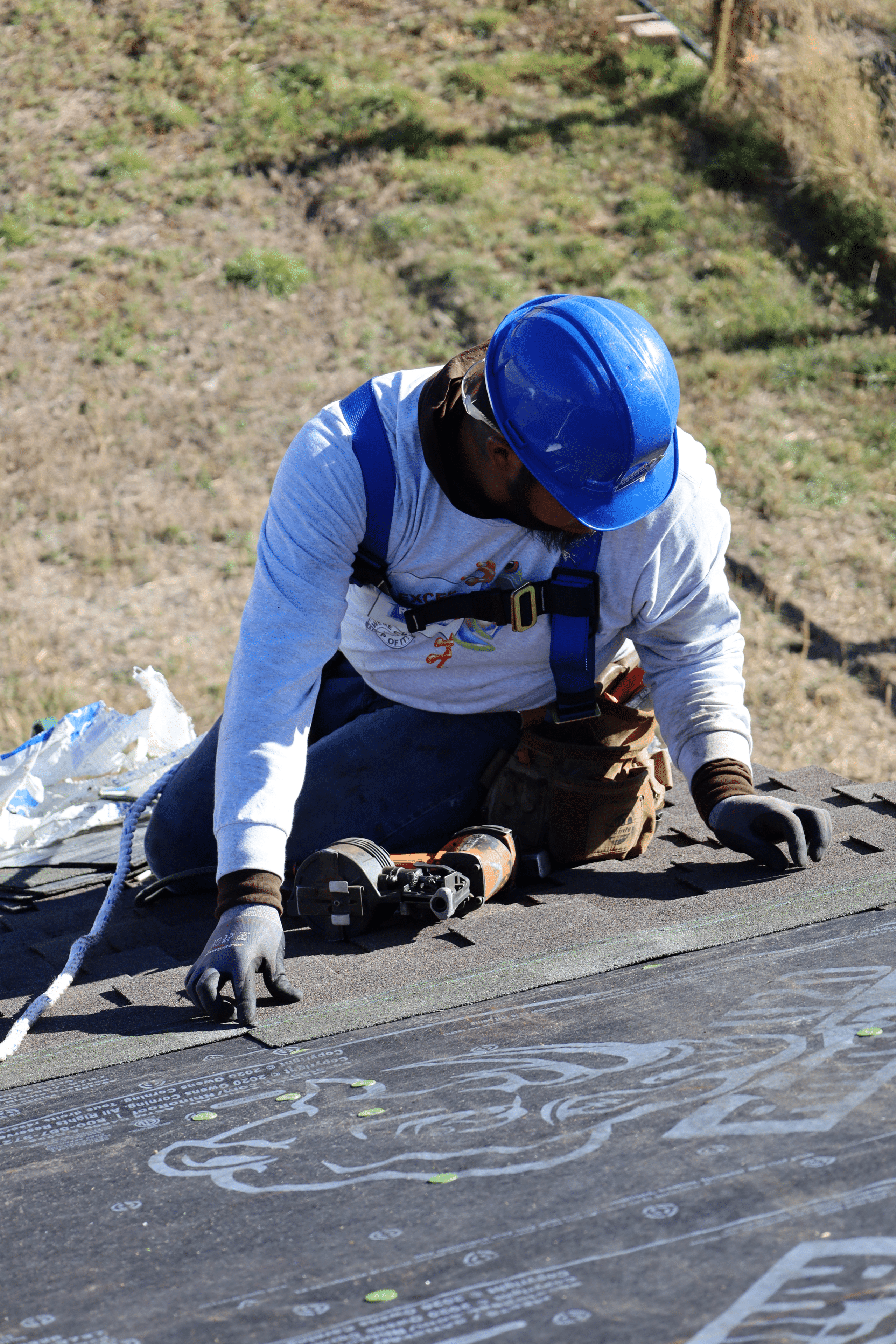 Man with a blue hard hat and harness sitting on roof, inspecting the work he has done.