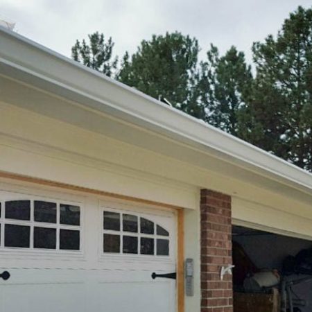 White rain gutters on a home placed above the garage. A drain pipe is seen going to the ground on one side. The left garage door is white and has sectioned windows at the top. The right garage door is open.