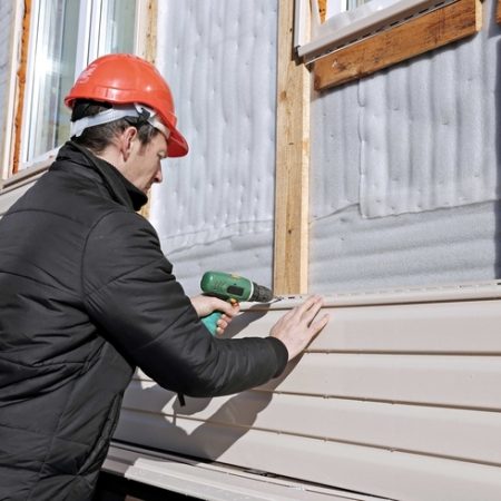 A kneeled worker wearing a red hard hat and a black jacket while installing tan siding on a house.